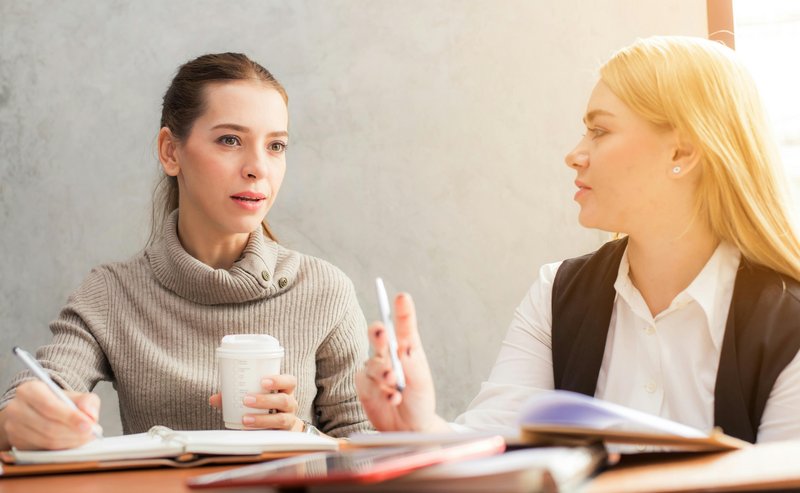Zwei Frauen sitzen nebeneinander an einem Konferenztisch und tauschen sich während eines Meetings freundlich aus.