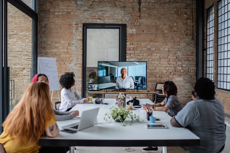 meeting-teilnehmer.jpg In einem modernen Besprechungsraum sitzen vier Geschäftspersonen an einem Tisch und führen ein hybrides Meeting mit einer weiteren Person durch, die per Videokonferenz auf einem Laptop zugeschaltet ist.